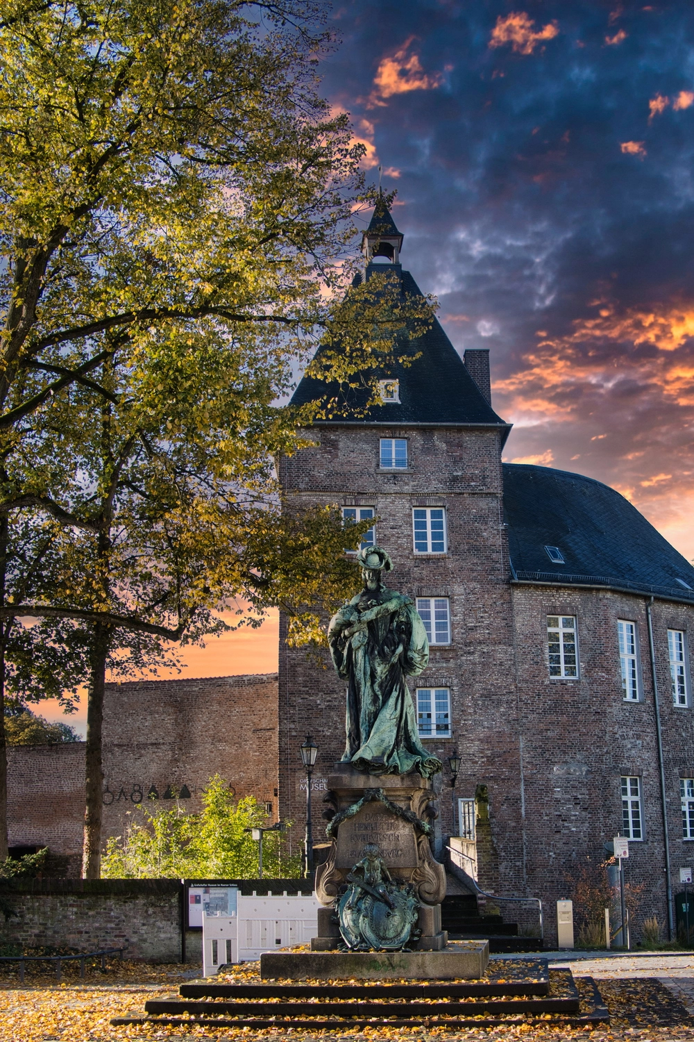 Rechtsanwaltskanzlei Paul - Mietrecht, Arbeitsrecht und Verkehrsrecht - Ein hoher Burgturm aus Backstein unter einem dramatischen Sonnenuntergangshimmel, im Vordergrund auf einem Platz steht eine grüne Bronzestatue auf einem Steinsockel.
